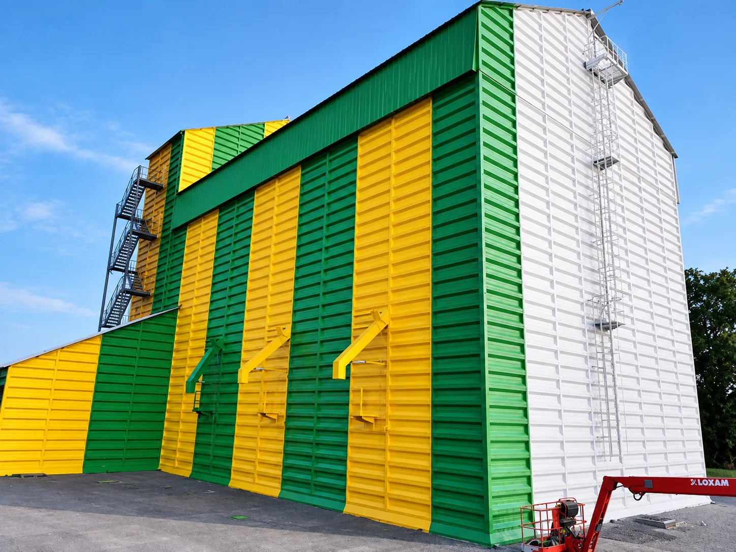 Silo peint en bandes verticales vert et jaune, pignon blanc, nacelle au pied du bâtiment.